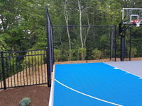 Blue and gray residential basketball court in Easton, MA, focused on left side of court, fence and goal.