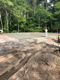 Similar view from finished picture to left, but bare ground in foreground and form with fresh cement being smoothed in to create a base for blue and gray residential basketball court in Easton, MA.