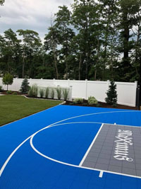 Side view of blue and gray residential basketball court in Easton, MA, highlighting landscaping by Evergreen beyond other side of court.