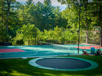 In-ground trampoline in foreground, installed along with construction of this large backyard multicourt, combining basketball, lines for secondary games, and an adjustable net for tennis or volleyball, located in Pembroke, MA.