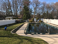 View from ground level, with entire pool covered by flooded tarp in foreground, before any tree removal or other prep was done for royal blue and yellow basketball court and accessories in Stoneham, MA, viewed from adjacent covered pool.