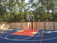 Finished picture of bulk of charcoal and orange home basketball court, looking toward hoop, showing part of custom fence that incorporates traditional cedar wood with rebounder mesh segments, in Walpole, MA.