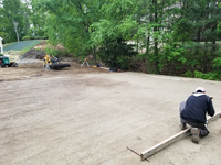Worker prepares form for cement to go on packed sand to build foundation of black and grey home backyard basketball court in Wellesley, MA.