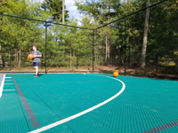 Low view showing closer view of green court tiles, with youngster shooting hoops in background, and view of optional fencing on backyard basketball court in Dover, MA.