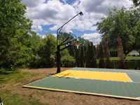 Angled left side view of olvie and yellow Easton, MA court in daylight, with lighting system visible above hoop.