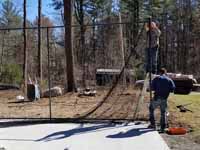Residential basketball court in Norwell, MA, including goal system, mesh fencing, and an emerald green and rust red tile sport surface. Some trees and stumps were removed to make room in the yard and provide easy work access. This shows workers installing net fencing onto previously installed posts.