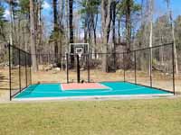 Residential basketball court in Norwell, MA, including goal system, mesh fencing, and an emerald green and rust red tile sport surface. Some trees and stumps were removed to make room in the yard and provide easy work access.