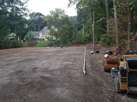Packing gravel underlay and the forms and reinforcement for concrete partially in place on the way to building a large emerald green and titanium backyard basketball court in Bolton, MA.