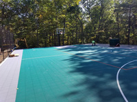 View of much of large emerald green and titanium backyard basketball court in Bolton, MA with tiles still being installed along one side.