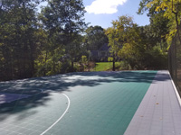 Sun-drenched long view of most of large emerald green and titanium backyard basketball court in Bolton, MA.